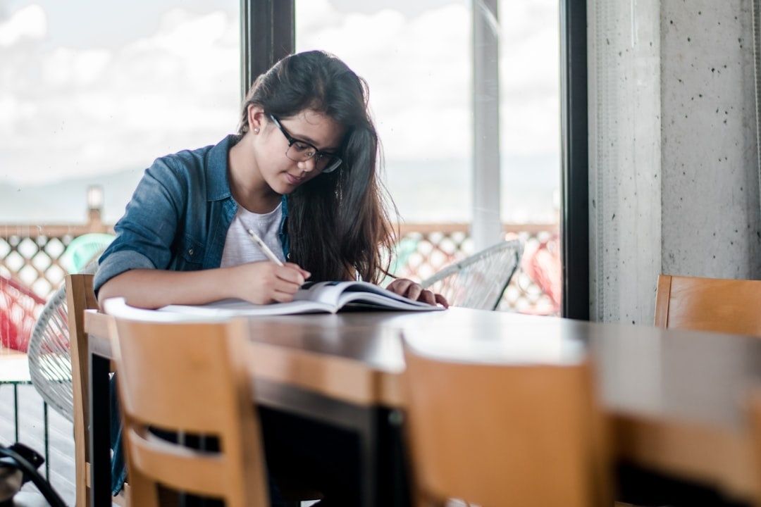 Student med glasögon skriver i en öppen bok vid ett bord